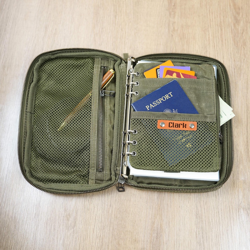 May include: Open olive green travel organizer with multiple compartments. Includes a mesh pocket, zippered pocket, and ring binder. Contains a pen, passport, cards, and a tag that says "Clark".