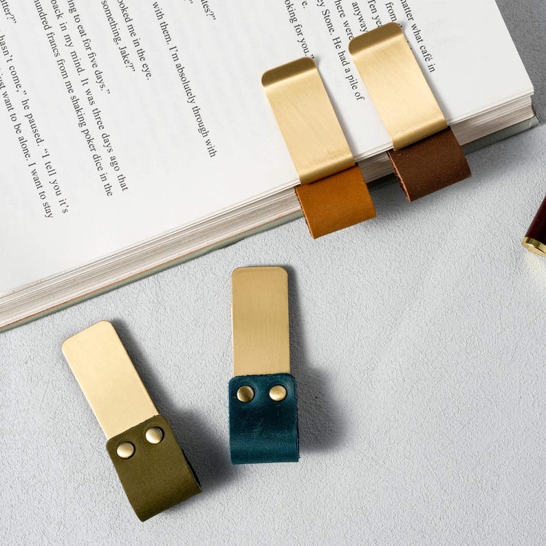 May include: A collection of brass and leather bookmarks in various colors, including brown, green, and teal. The bookmarks are rectangular with a brass top and a leather strap. Some are placed on the pages of a book.