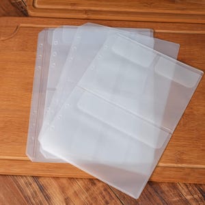 May include: A stack of clear, plastic binder sleeves with pockets and pre-punched holes for organization. The sleeves are translucent, allowing visibility of contents. The image is taken on a wooden surface.