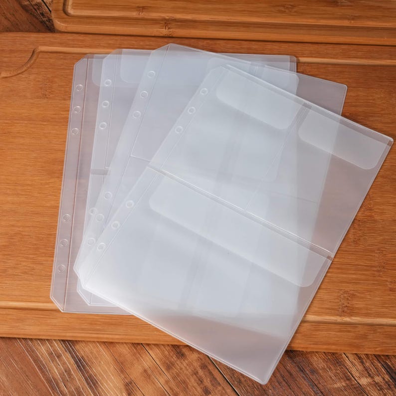 May include: A stack of clear, plastic binder sleeves with pockets and pre-punched holes for organization. The sleeves are translucent, allowing visibility of contents. The image is taken on a wooden surface.