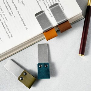 May include: A collection of metal and leather bookmarks in various colors, including brown, orange, and teal. The bookmarks are rectangular with a metal top and a leather bottom, attached with rivets. A pen rests on the side.