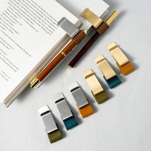 May include: A collection of pen holders and page markers. The pen holders feature a combination of wood, metal, and leather. The page markers come in various colors, including gold, silver, green, and orange. The items are arranged on a book.