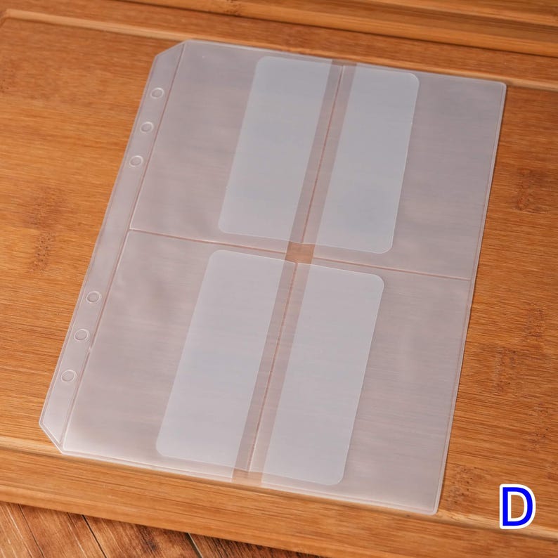 May include: A clear plastic card holder with four pockets, each designed to hold a standard-sized card. The holder has a row of holes along the left edge for binding and is set against a wooden surface. The letter "D" is in the bottom right corner.