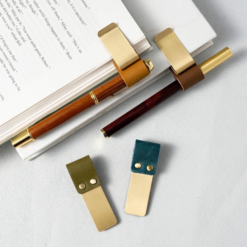 May include: Two pens with brass clips and leather accents rest on a book. Two leather and brass bookmarks are also visible. One pen is brown, the other is dark red. The bookmarks are green and blue.