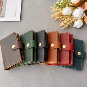 May include: Six leather-bound notebooks in different colors, including brown, green, black, orange, red, and dark blue. The notebooks have a gold button closure and are arranged in a row.