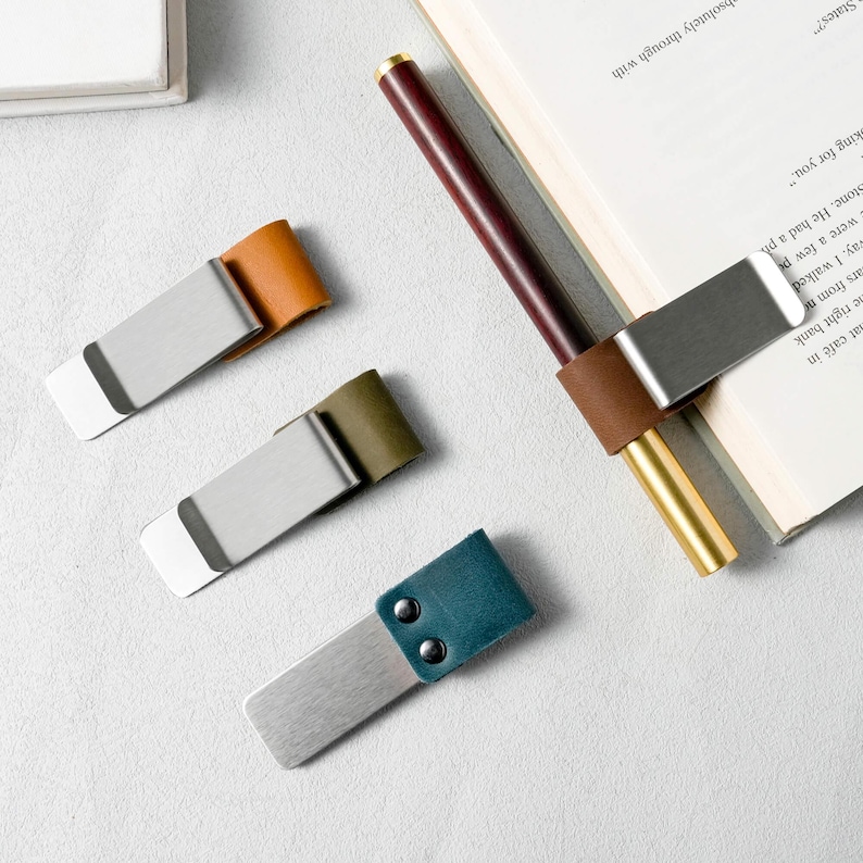 May include: A collection of metal and leather bookmarks in various colors, including tan, olive green, and teal. One bookmark is attached to a pen and a book. The bookmarks have a minimalist design.