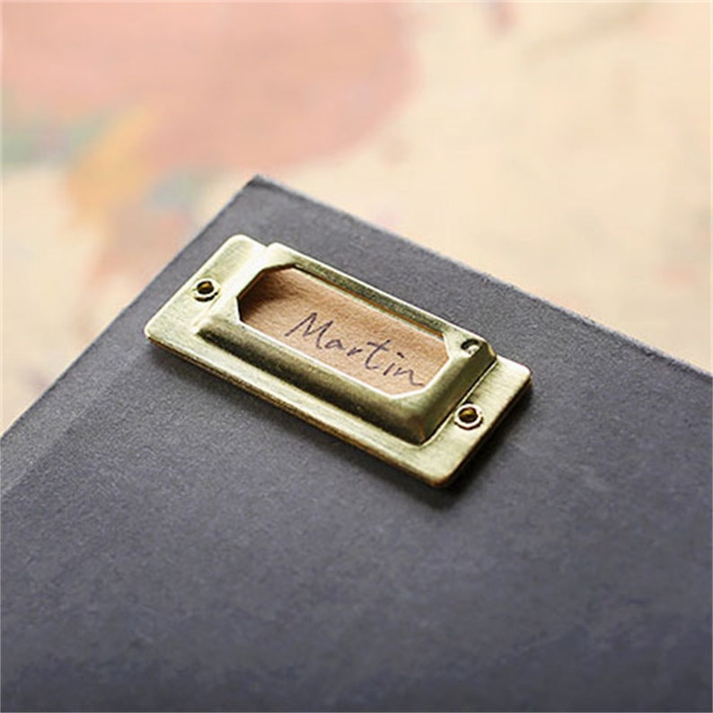 May include: A gold metal label holder with a rectangular shape and rounded corners. The label holder has a small wooden insert with the name "Martin" written on it.