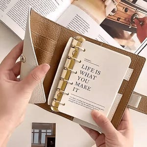 May include: A white and brown leather planner with gold-colored rings, open to a page that reads "LIFE IS WHAT YOU MAKE IT." The planner is held open, revealing a glimpse of a magazine in the background.