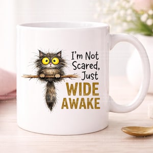 May include: White ceramic mug featuring a whimsical illustration of a wide-eyed, fluffy cat perched on a branch. The mug has the text "I'm Not Scared, Just WIDE AWAKE" in black and gold lettering. A fun, novelty item.