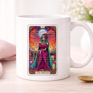 Puede incluir: Una taza de cer&aacute;mica blanca con una colorida ilustraci&oacute;n de carta de tarot titulada THE SPIRITED. La imagen muestra una figura esquel&eacute;tica con cabello verde azulado en un vestido magenta, sosteniendo una copa frente a un cielo al atardecer.