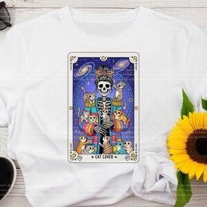 Puede incluir: Una camiseta blanca con un gr&aacute;fico de un esqueleto con un mo&ntilde;o que lleva un c&aacute;rdigan de rayas de colores, rodeado de varios gatos peque&ntilde;os. El dise&ntilde;o est&aacute; enmarcado como una carta del tarot sobre un fondo azul oscuro estrellado.