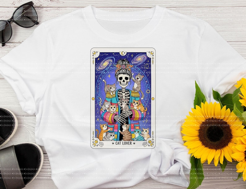 Puede incluir: Una camiseta blanca con un gr&aacute;fico de un esqueleto con un mo&ntilde;o que lleva un c&aacute;rdigan de rayas de colores, rodeado de varios gatos peque&ntilde;os. El dise&ntilde;o est&aacute; enmarcado como una carta del tarot sobre un fondo azul oscuro estrellado.
