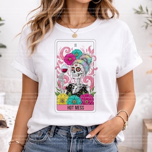 Puede incluir: Una camiseta blanca de manga corta con un gr&aacute;fico de un esqueleto con un mo&ntilde;o de colores, sosteniendo una copa de vino tinto. El dise&ntilde;o est&aacute; enmarcado como una carta del tarot con detalles florales en rosa, amarillo y turquesa, y el texto HOT MESS.