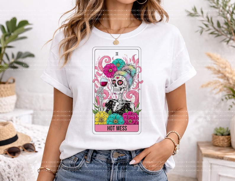 Puede incluir: Una camiseta blanca de manga corta con un gr&aacute;fico de un esqueleto con un mo&ntilde;o de colores, sosteniendo una copa de vino tinto. El dise&ntilde;o est&aacute; enmarcado como una carta del tarot con detalles florales en rosa, amarillo y turquesa, y el texto HOT MESS.