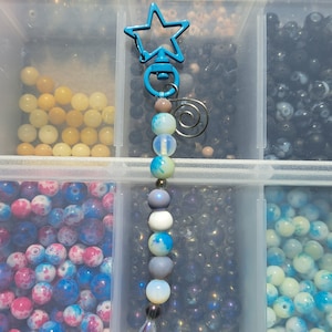Puede incluir: Un llavero de cuentas con un dije en forma de estrella azul. El llavero tiene una variedad de cuentas en tonos azules, grises y blancos. Las cuentas están dispuestas en una línea vertical y se intercalan con cuentas transparentes. El llavero está colgado de un recipiente de plástico transparente lleno de cuentas.