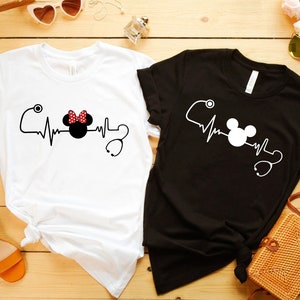 May include: Two t-shirts with a heartbeat graphic featuring Mickey Mouse and Minnie Mouse with a stethoscope. The white t-shirt has Minnie Mouse and the black t-shirt has Mickey Mouse.