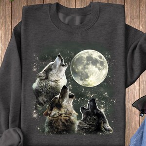K&ouml;nnte beinhalten: Ein dunkelgraues Sweatshirt mit einem grafischen Druck von drei W&ouml;lfen, die zum Mond heulen. Die W&ouml;lfe sind grau und wei&szlig;, und der Mond ist leuchtend gelb.
