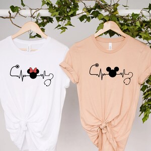 May include: Two t-shirts with a heartbeat graphic featuring a Mickey Mouse head and a Minnie Mouse head with a red polka dot bow. The t-shirts are hanging on hangers against a white background.