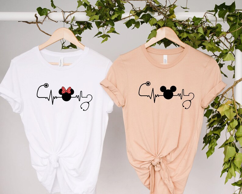 May include: Two t-shirts with a heartbeat graphic featuring a Mickey Mouse head and a Minnie Mouse head with a red polka dot bow. The t-shirts are hanging on hangers against a white background.