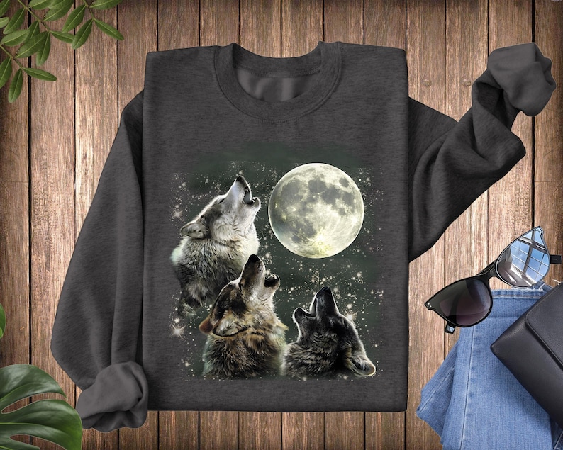 K&ouml;nnte beinhalten: Ein dunkelgraues Sweatshirt mit einem grafischen Druck von drei W&ouml;lfen, die zum Mond heulen. Die W&ouml;lfe sind grau und wei&szlig;, und der Mond ist leuchtend gelb.