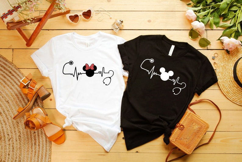 May include: Two t-shirts with a heartbeat graphic featuring Mickey Mouse and Minnie Mouse with a stethoscope. The white t-shirt has Minnie Mouse and the black t-shirt has Mickey Mouse.