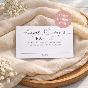 Puede incluir: Una tarjeta blanca con la inscripción en negro "diaper & wipes raffle", que invita a los invitados a traer pañales o toallitas húmedas para un premio. La tarjeta está sobre una bandeja de madera con una tela beige y detalles florales.