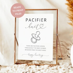 May include: A framed white sign with the words "PACIFIER hunt" and a pacifier illustration. The sign also includes the text "THERE ARE PACIFIERS HIDDEN THROUGHOUT THE BABY SHOWER. WHOEVER FINDS THE MOST WINS! happy hunting."