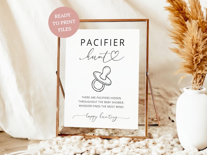 K&ouml;nnte beinhalten: Ein wei&szlig;es rechteckiges Schild in einem kupferfarbenen Rahmen mit dem schwarzen Text PACIFIER hunt, einem Herzsymbol, einer Strichzeichnung eines Schnullers und dem Text THERE ARE PACIFIERS HIDDEN THROUGHOUT THE BABY SHOWER. WHOEVER FINDS THE MOST WINS! happy hunting.