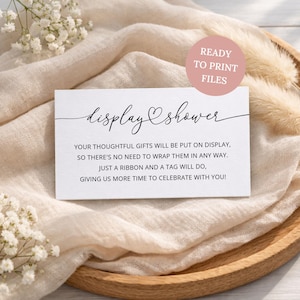 Op de afbeelding: Een rechthoekige witte kaart met zwarte tekst die luidt display shower, your thoughtful gifts will be put on display, so there's no need to wrap them in any way. Just a ribbon and a tag will do, giving us more time to celebrate with you!