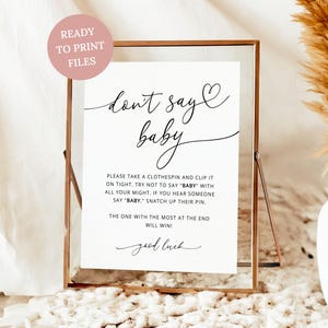 May include: A framed white sign with the text "don't say baby" in elegant script. The sign includes instructions for a baby shower game. A pink circle with the text "READY TO PRINT FILES" is in the upper left corner.