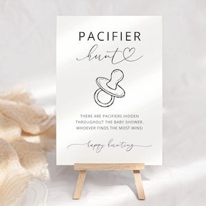 K&ouml;nnte beinhalten: Ein wei&szlig;es rechteckiges Schild mit schwarzem Text und einer Strichzeichnung eines Schnullers. Der Text lautet: PACIFIER hunt, THERE ARE PACIFIERS HIDDEN THROUGHOUT THE BABY SHOWER. WHOEVER FINDS THE MOST WINS!, happy hunting. Das Schild steht auf einer Staffelei.