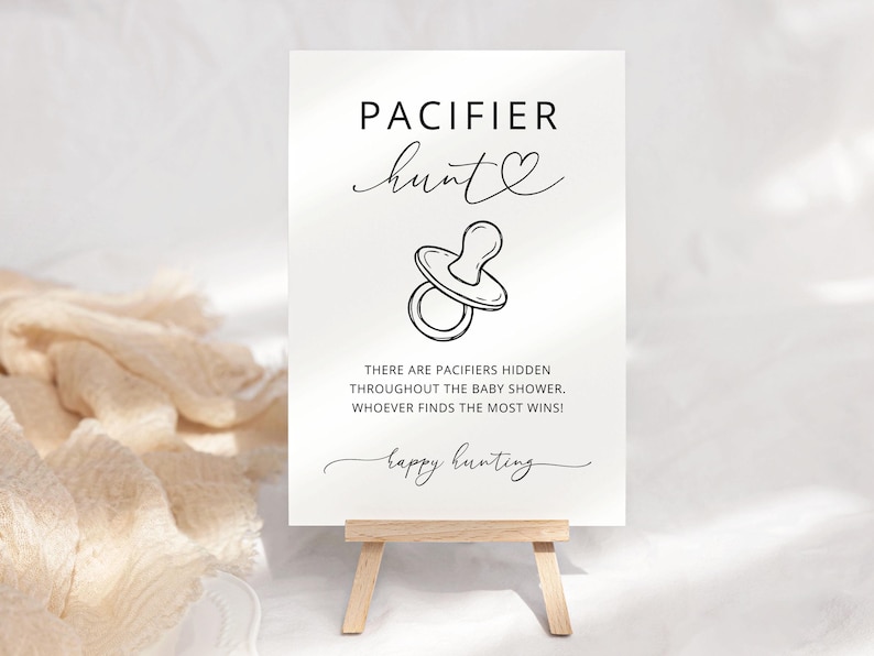 K&ouml;nnte beinhalten: Ein wei&szlig;es rechteckiges Schild mit schwarzem Text und einer Strichzeichnung eines Schnullers. Der Text lautet: PACIFIER hunt, THERE ARE PACIFIERS HIDDEN THROUGHOUT THE BABY SHOWER. WHOEVER FINDS THE MOST WINS!, happy hunting. Das Schild steht auf einer Staffelei.