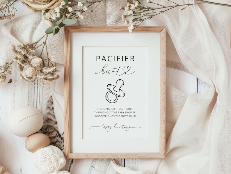 K&ouml;nnte beinhalten: Ein rechteckiger Holzrahmen mit einem wei&szlig;en Schild mit schwarzem Text, der PACIFIER hunt lautet, einer Strichzeichnung eines Schnullers und der Anweisung: THERE ARE PACIFIERS HIDDEN THROUGHOUT THE BABY SHOWER. WHOEVER FINDS THE MOST WINS! gefolgt von happy hunting.