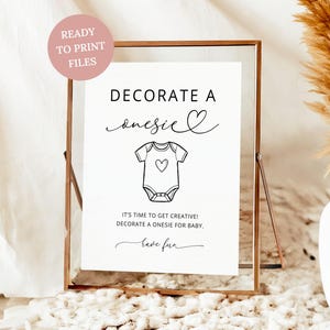May include: A framed print with the text "DECORATE A onesie" and a onesie illustration with a heart. The print also includes the text "IT'S TIME TO GET CREATIVE! DECORATE A ONESIE FOR BABY. have fun."