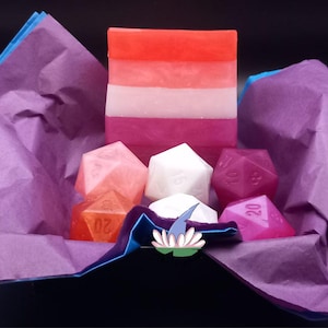 Op de afbeelding: Een geschenkdoos met paars vloeipapier bevat een gelaagde zeep in tinten rood, roze en wit, samen met verschillende veelvlakkige dobbelstenen in verschillende kleuren. Er is ook een kleine, kleurrijke speld zichtbaar.