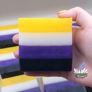 Puede incluir: Una pastilla de jabón cuadrada con el diseño de la bandera del orgullo no binario. El jabón presenta rayas horizontales amarillas, blancas, moradas y negras. Se ven otras pastillas de jabón en el fondo. El jabón se sostiene en una mano.