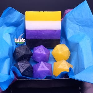 Puede incluir: Una caja de regalo con papel de seda azul contiene una pastilla de jabón con los colores de la bandera del orgullo no binario y varios jabones con forma de dados en negro, morado y amarillo. La pastilla de jabón es amarilla, blanca, morada y negra.