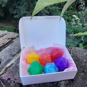Könnte beinhalten: Ein Set mit sechs bunten Würfeln in einer weißen Box mit rosa Seidenpapier. Die Würfel haben die Form eines zwanzigseitigen Würfels und sind rot, orange, gelb, grün, blau und lila.