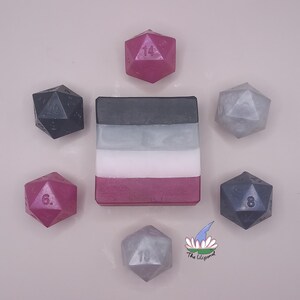 Op de afbeelding: Een handgemaakte zeep in de kleuren van de aseksuele vlag: zwart, grijs, wit en paars. De zeep is omringd door zeven twintigzijdige dobbelstenen in verschillende kleuren, waaronder roze, grijs en zwart. Het Lilipond-logo staat rechtsonder.