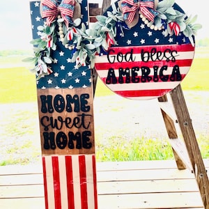 May include: Two patriotic wooden signs. One is rectangular with a blue star field, the words "HOME SWEET HOME", and red and white stripes. The other is round with red and white stripes and the words "God bless AMERICA". Both have bows and greenery.