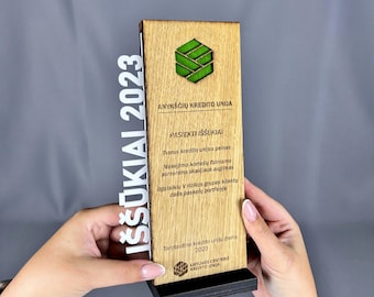 May include: A wooden award with the text "IŠŠŪKIAI 2023" in white. The award features a green geometric logo and text in Lithuanian. The base is black, and the main body is light brown.