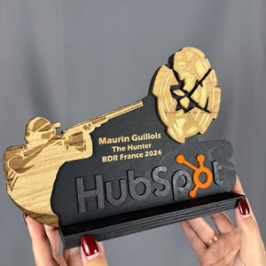 Puede incluir: Una placa de premio de madera con fondo negro y una silueta dorada y marrón de una persona sosteniendo una escopeta. El texto "Maurin Guillois The Hunter BDR France 2024" está impreso en dorado en la placa. La palabra "HubSpot" está impresa en negro con un logotipo naranja.