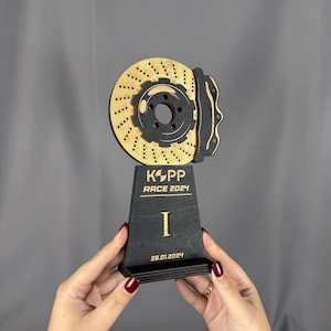 Peut inclure: Un trophée en bois noir et or avec un design de disque de frein. Le trophée porte les inscriptions "KOPP RACE 2024" et "I". La date "26.01.2024" est inscrite sur le socle.