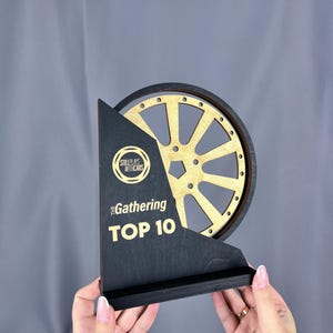 May include: A wooden trophy with a black base and a gold-colored wheel design. The trophy has the text "STILLPLAYS WITH CARS" and "The Gathering TOP 10" in gold lettering. The trophy is held up.
