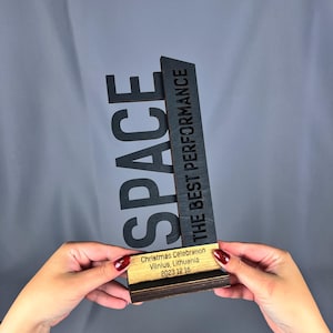 Op de afbeelding: Een zwarte houten award met de tekst "SPACE" en "THE BEST PERFORMANCE" erop. De award heeft een klein houten plaatje met de tekst "Christmas Celebration Vilnius, Lithuania 2023 12 16".