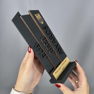 May include: A black wooden award plaque with gold lettering. The plaque reads "FHA Beer Awards" and "Pale Lager of the Year 2024".