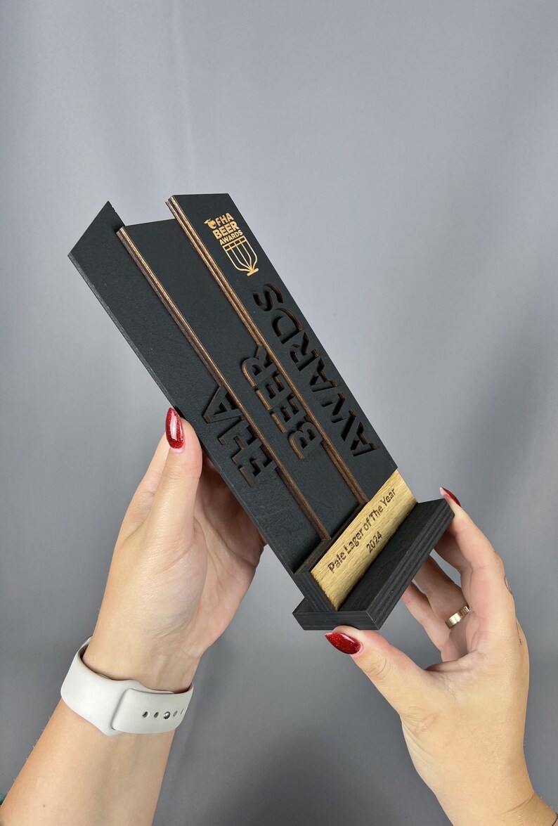 May include: A black wooden award plaque with gold lettering. The plaque reads "FHA Beer Awards" and "Pale Lager of the Year 2024".