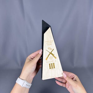Puede incluir: Trofeo de madera con parte superior negra y frontal triangular claro. El frente presenta texto dorado y rifles cruzados. El texto dice "LKMEY 2022 II ETAPO TRUPE" y "GRUPE-A III".