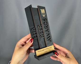 May include: Black wooden award plaque with gold lettering for the "FHA Beer Awards". The plaque has a cutout of a beer glass and the words "FHA Beer Awards". The plaque has a wooden base with the text "Pale Lager of the Year 2024".