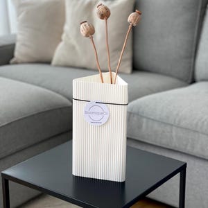 Puede incluir: Jarrón de cerámica blanco con nervaduras y una pegatina redonda que dice "BloomiqueCo" sobre una mesa auxiliar negra. Hay cápsulas de semillas de amapola secas en el jarrón.